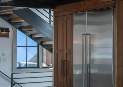 An architectural view full of angles, metal and wood treat the eye like a geometric art rather than a view of the staircase, windows, kitchen cabinetry, and a refrigerator.